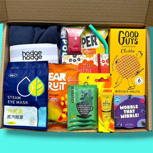A curated gift box filled with assorted packaged snacks, tea, and wellness items, photographed from above inside an open cardboard box against a bright turquoise background. Visible items include a dark blue folded towel or cloth, a blue 'steam eye mask' packet, an orange 'ear fruit' packet, a green 'clipper' packet, a yellow 'Good Guys' biscuit melts box, a purple 'Wobble That Wibble!' packet, and other colorful snack and tea packages.