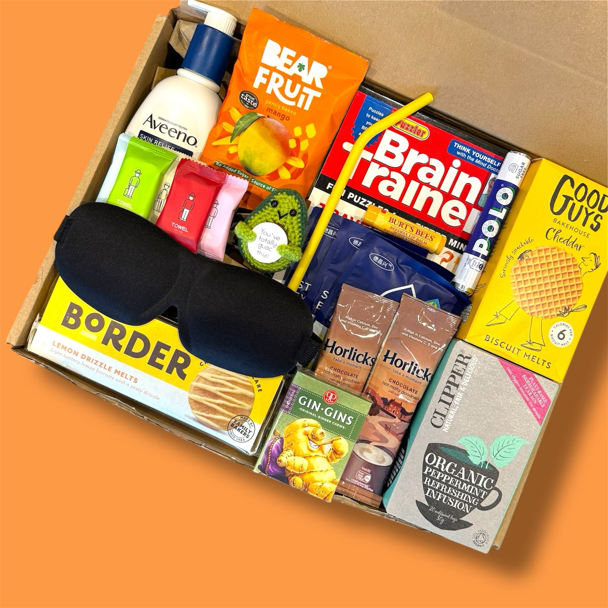 Assorted items including snacks, drinks, and a sleep mask in a box on an orange background