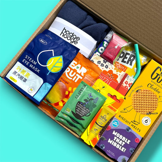 A curated gift box filled with assorted packaged snacks, tea, and a pair of dark navy socks, photographed from above on a bright aqua background.